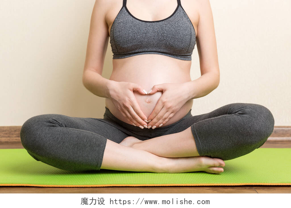一个孕妇坐在瑜伽垫上盘腿漂亮的孕妇在家做瑜伽。验尸时的妊娠期瑜珈与健康概念.